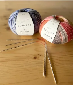 Deux pelotes de laine Katia Concept Aiti Socks et Heli Socks avec aiguilles circulaires sur une table en bois clair.